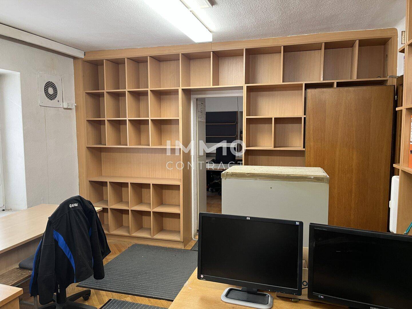 Büroraum mit großen Holzregalen, zwei Computermonitoren und einem Schreibtisch.