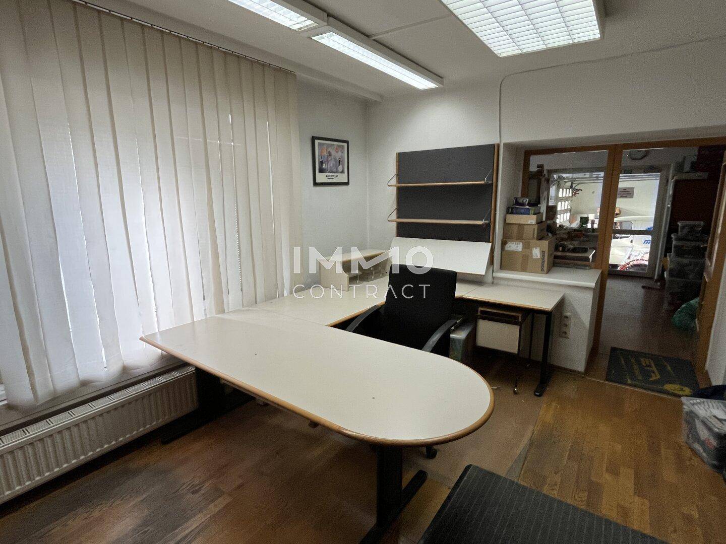 Büroraum mit Schreibtisch, Bürostuhl und vertikalen Jalousien am Fenster.