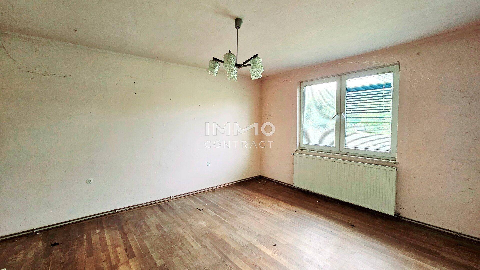 Leeres Zimmer mit abgenutztem Parkettboden, einem Fenster und einem Heizkörper in renovierungsbedürftigem Zustand.