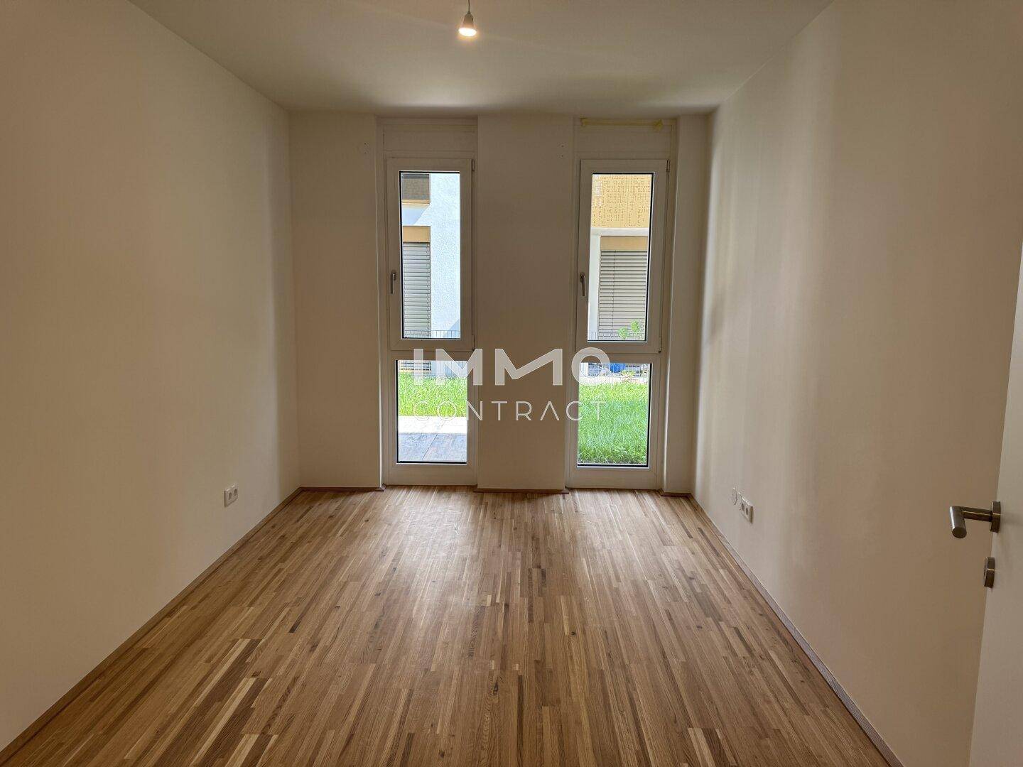 Lichtdurchfluteter Raum mit Holzboden und zwei großen Fenstertüren zum Außenbereich.
