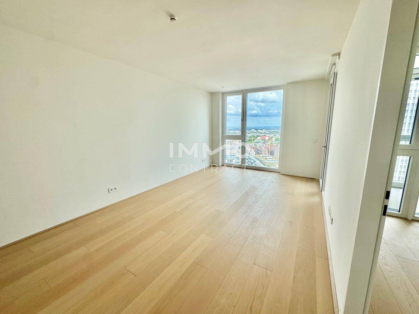 Heller, leerer Raum mit Holzboden und großen Fenstern, die einen weiten Stadtblick bieten.