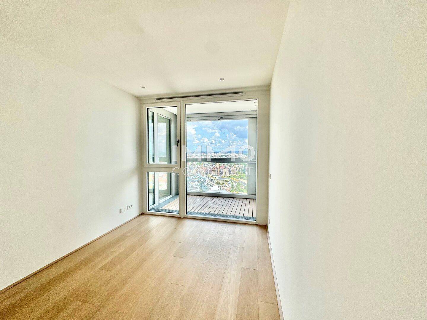 Leerer Raum mit Holzboden und Zugang zu einem Balkon, der einen weiten Blick über die Stadt ermöglicht.