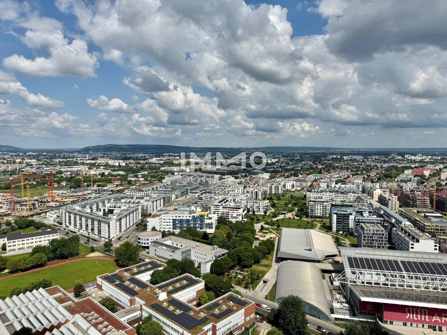 Weitläufige Luftaufnahme einer modernen Stadtlandschaft mit Wohngebäuden und Grünflächen unter blauem Himmel.