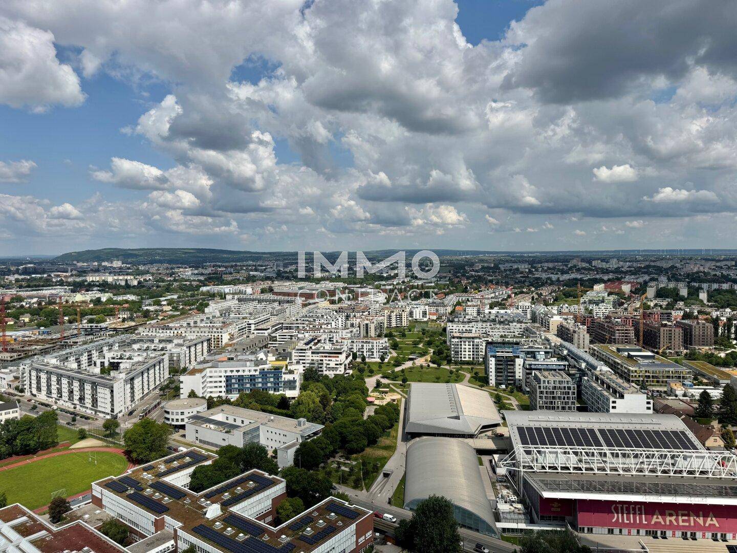 Weitläufiger Blick über eine städtische Landschaft mit vielen Gebäuden und Grünflächen unter blauem Himmel.