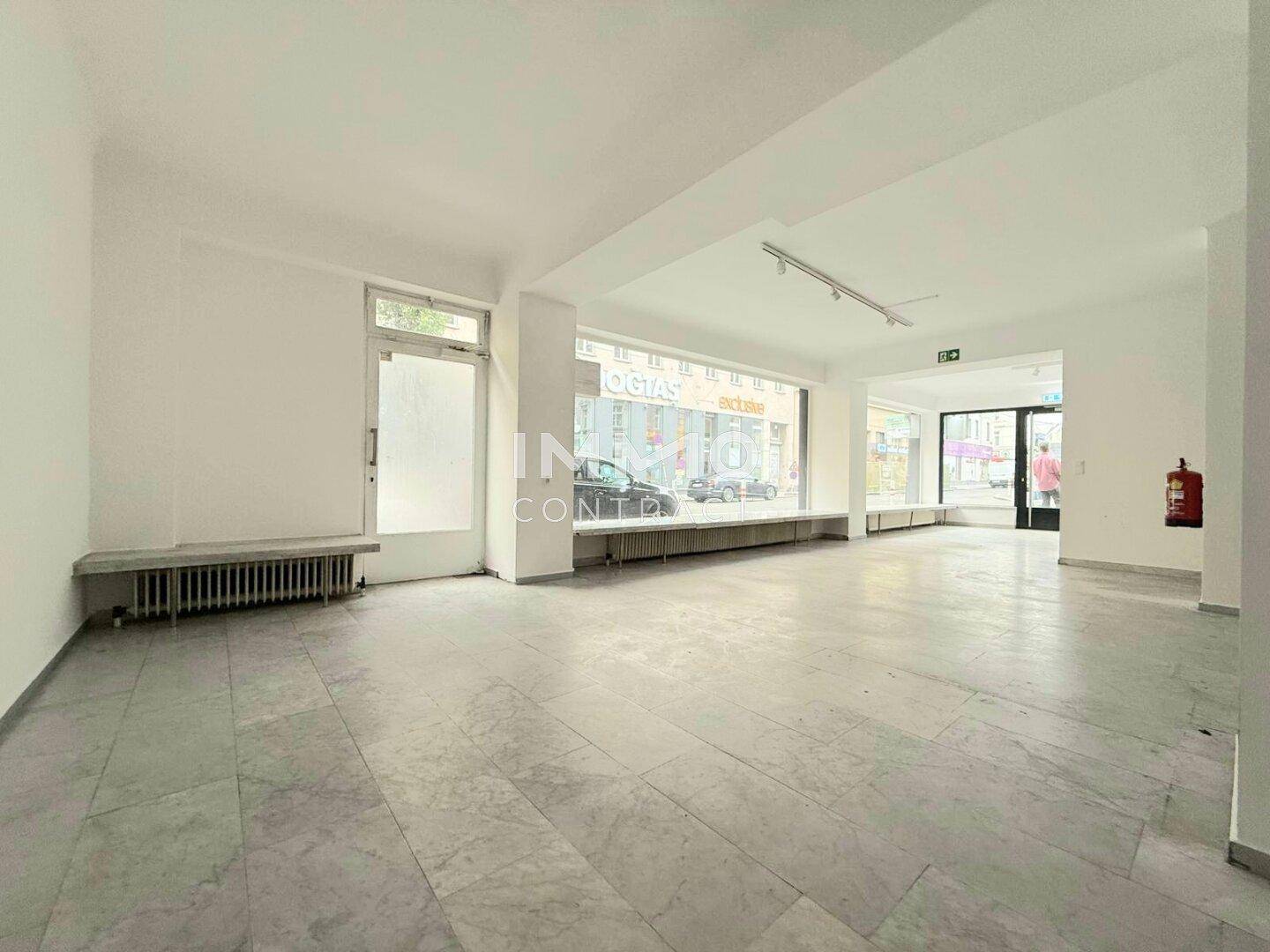 Großer, leerer Verkaufsraum mit Schaufenstern zur Straße und Heizkörpern unter den Fenstern.