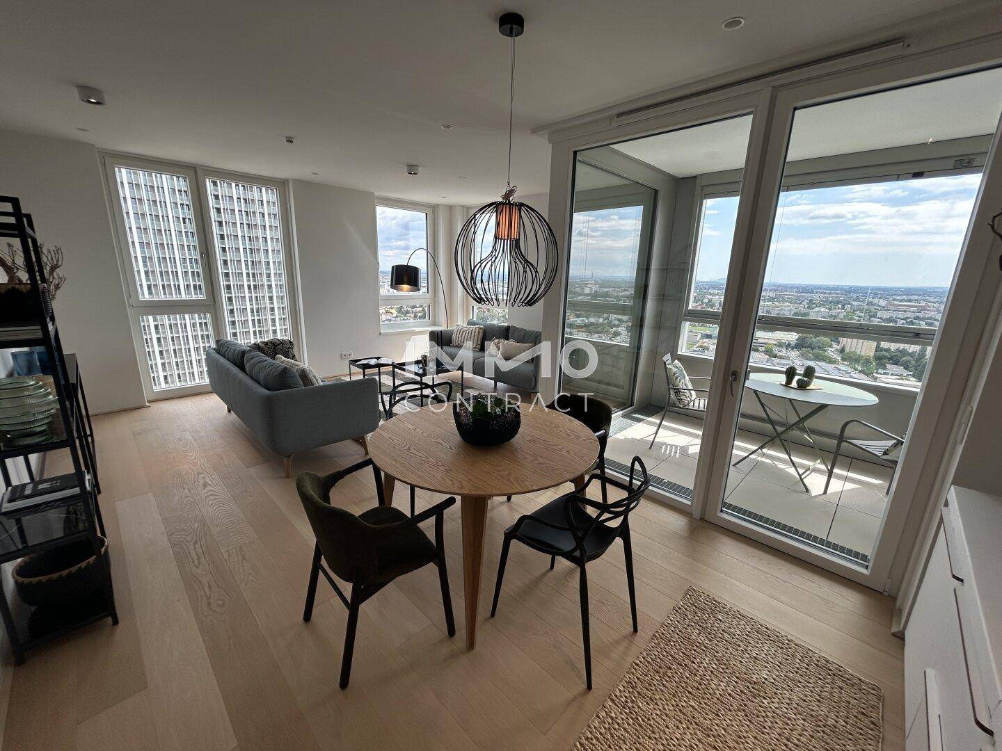 Lichtdurchfluteter Wohn-Essbereich mit Holzboden, moderner Einrichtung und Zugang zu einem verglasten Balkon mit Stadtblick.