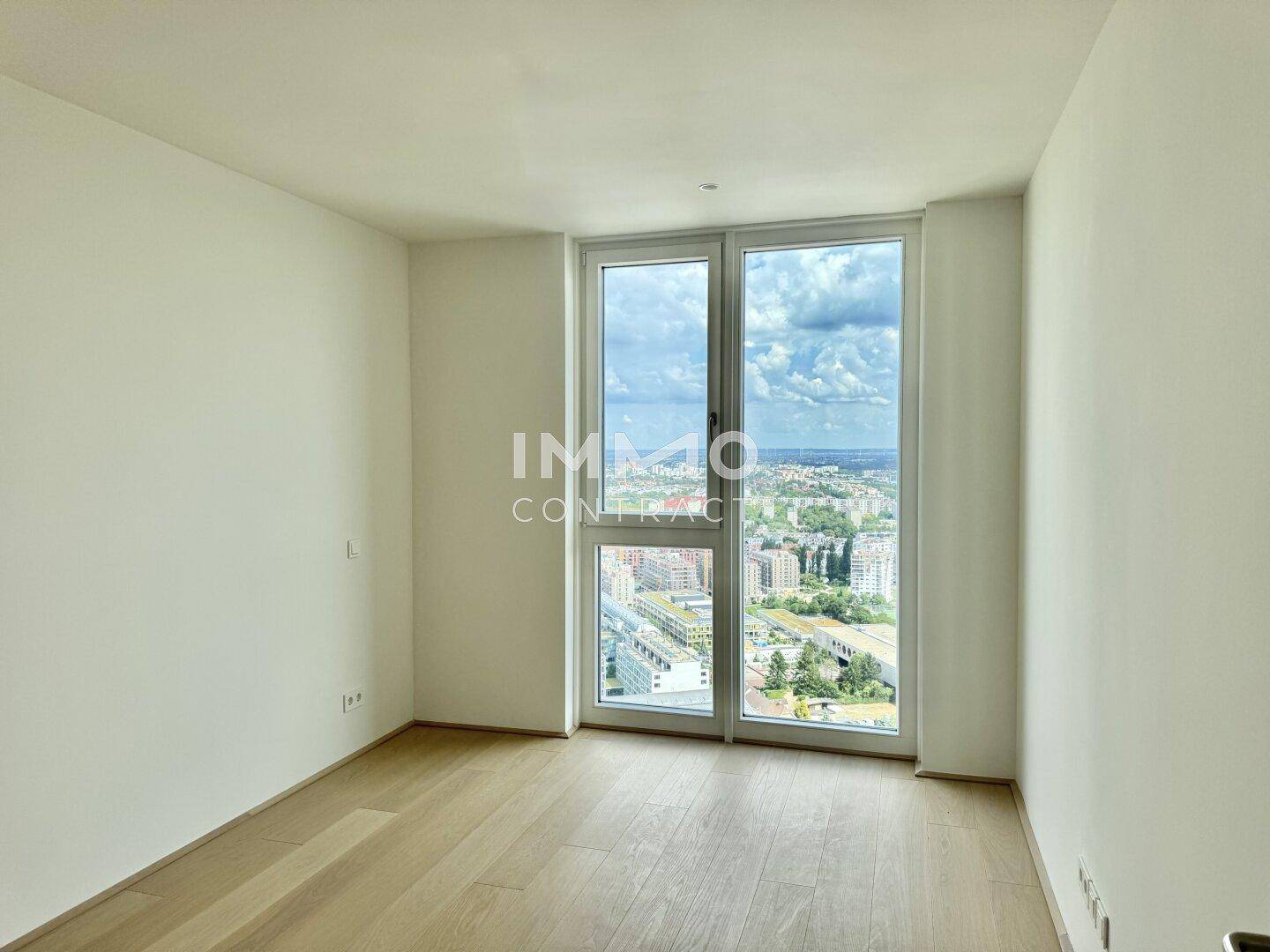 Helles, leeres Schlafzimmer mit Holzboden und bodentiefen Fenstern, die einen weiten Blick über die Stadt bieten.