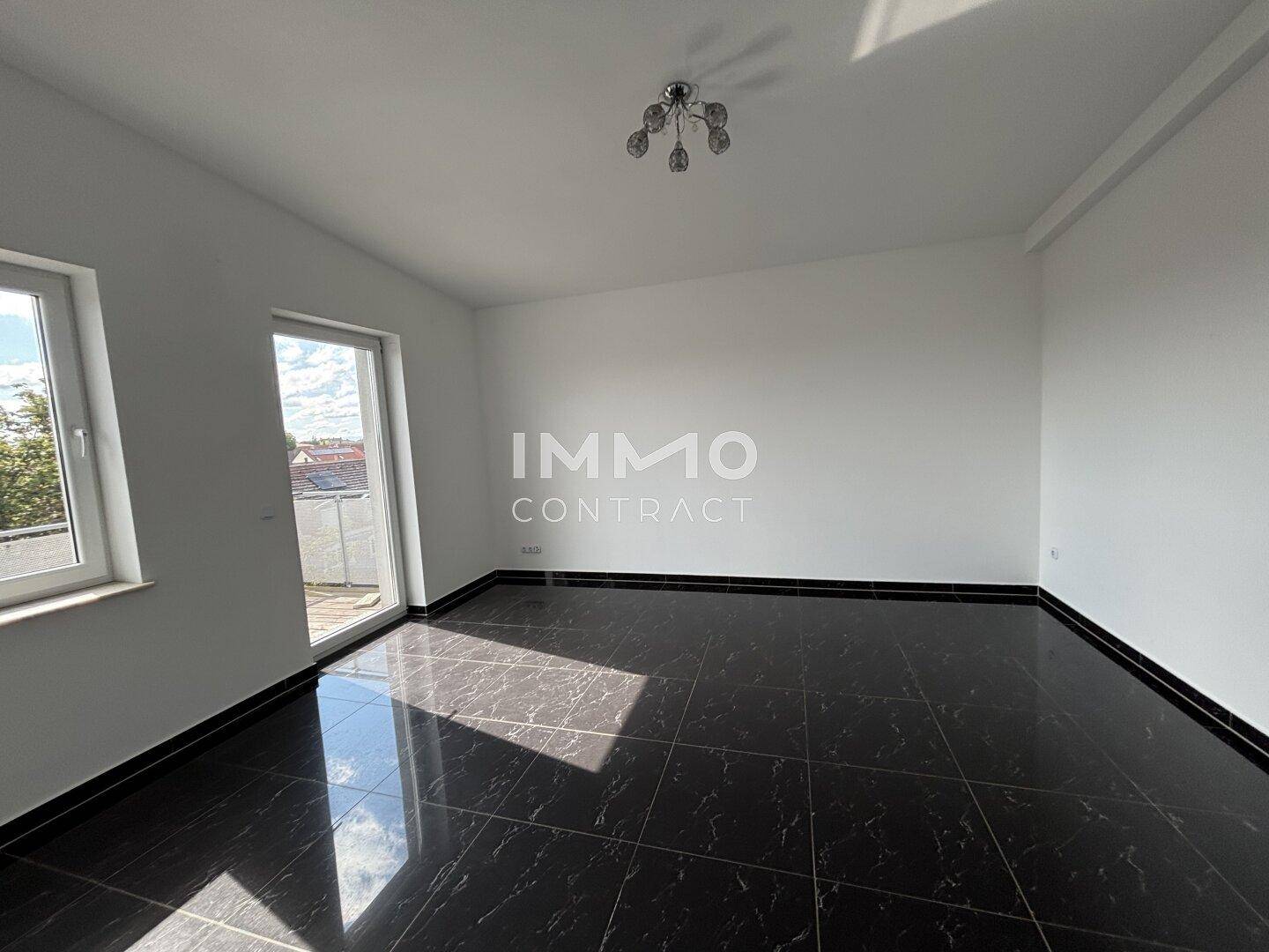 Heller, leerer Wohnraum mit glänzendem schwarzen Fliesenboden und Zugang zu einem Balkon.