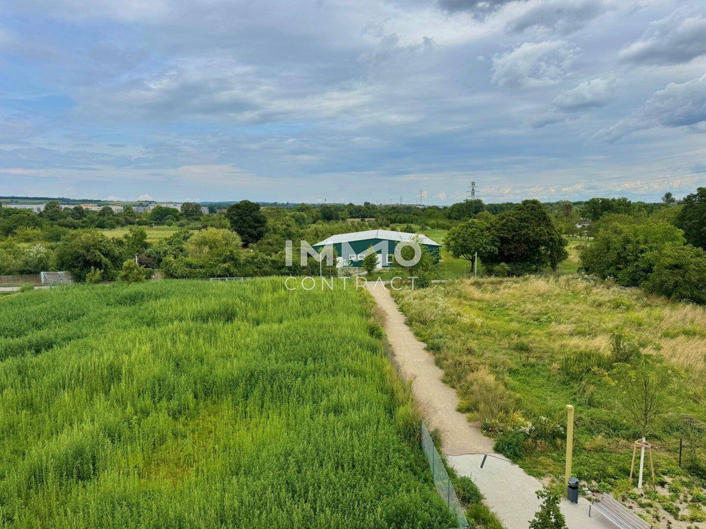 Weitläufiger Blick über grüne Felder und eine ländliche Umgebung mit einem Spazierweg.