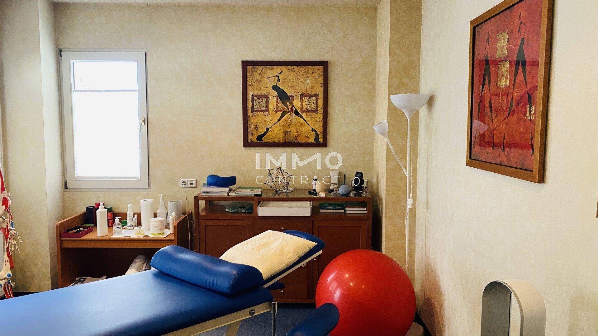 Ruhiger Therapieraum mit einer Behandlungsliege, einem Fenster und dekorativen Bildern an den Wänden.