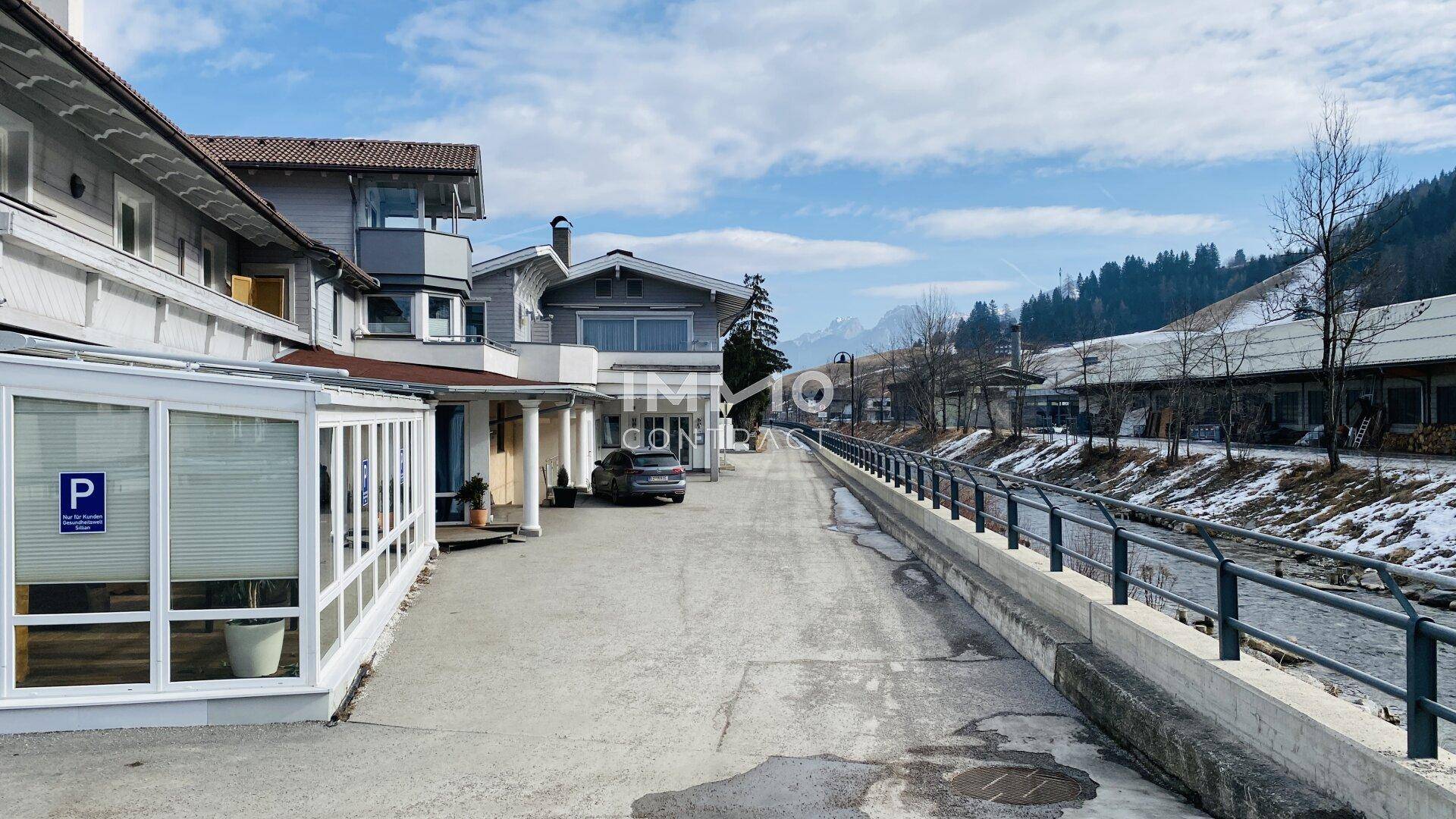 Winterliche Außenansicht eines Gebäudekomplexes mit Parkplätzen und einem Fluss im Vordergrund, umgeben von Bergen.