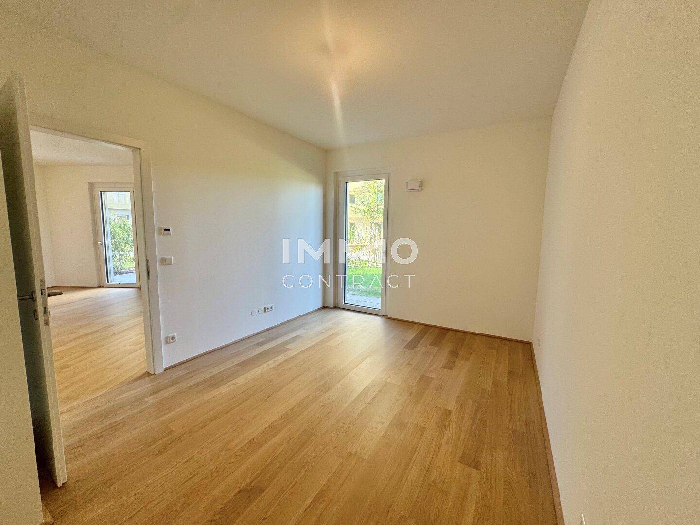 Leeres Zimmer mit hellem Parkettboden und einer Glastür, die viel natürliches Licht hereinlässt.