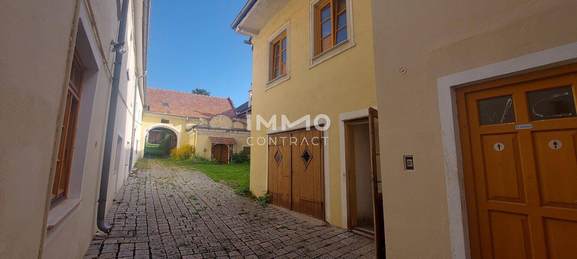 Schmaler gepflasterter Weg zwischen historischen Gebäuden, der zu einem sonnigen Innenhof führt.