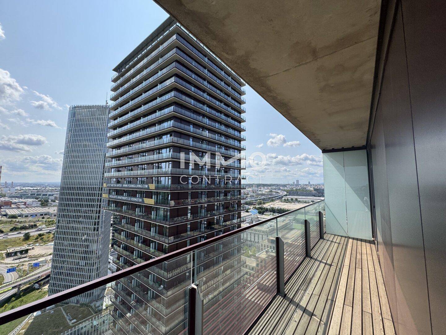 Balkon mit Holzboden und Glasgeländer, der einen Blick auf das moderne Hochhaus nebenan bietet.