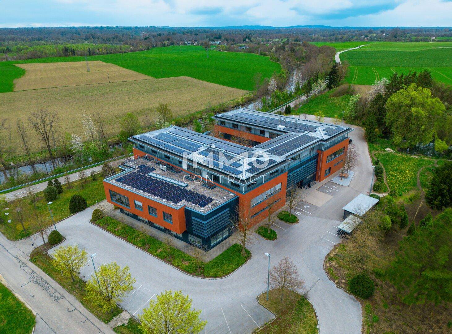 Panoramische Luftaufnahme eines modernen Bürogebäudes mit umfangreichen Solarmodulen, umgeben von Feldern, einem Fluss und Bäumen.