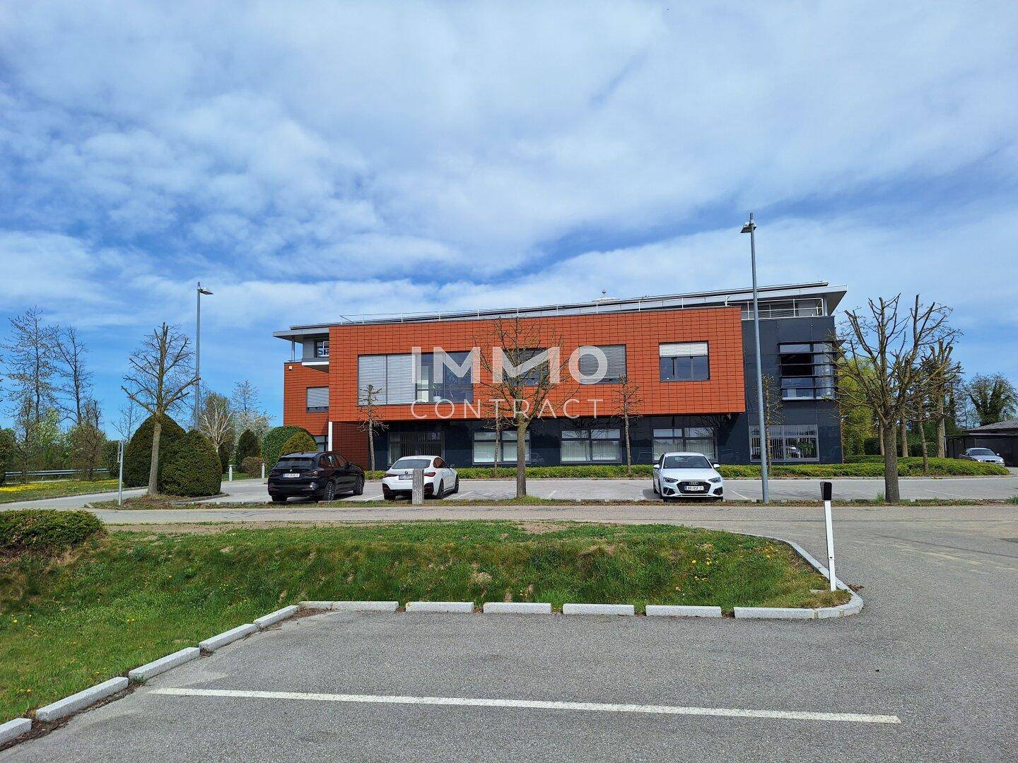 Modernes Bürogebäude mit orangefarbener und dunkelgrauer Fassade, umgeben von einem großen Parkplatz unter blauem Himmel.