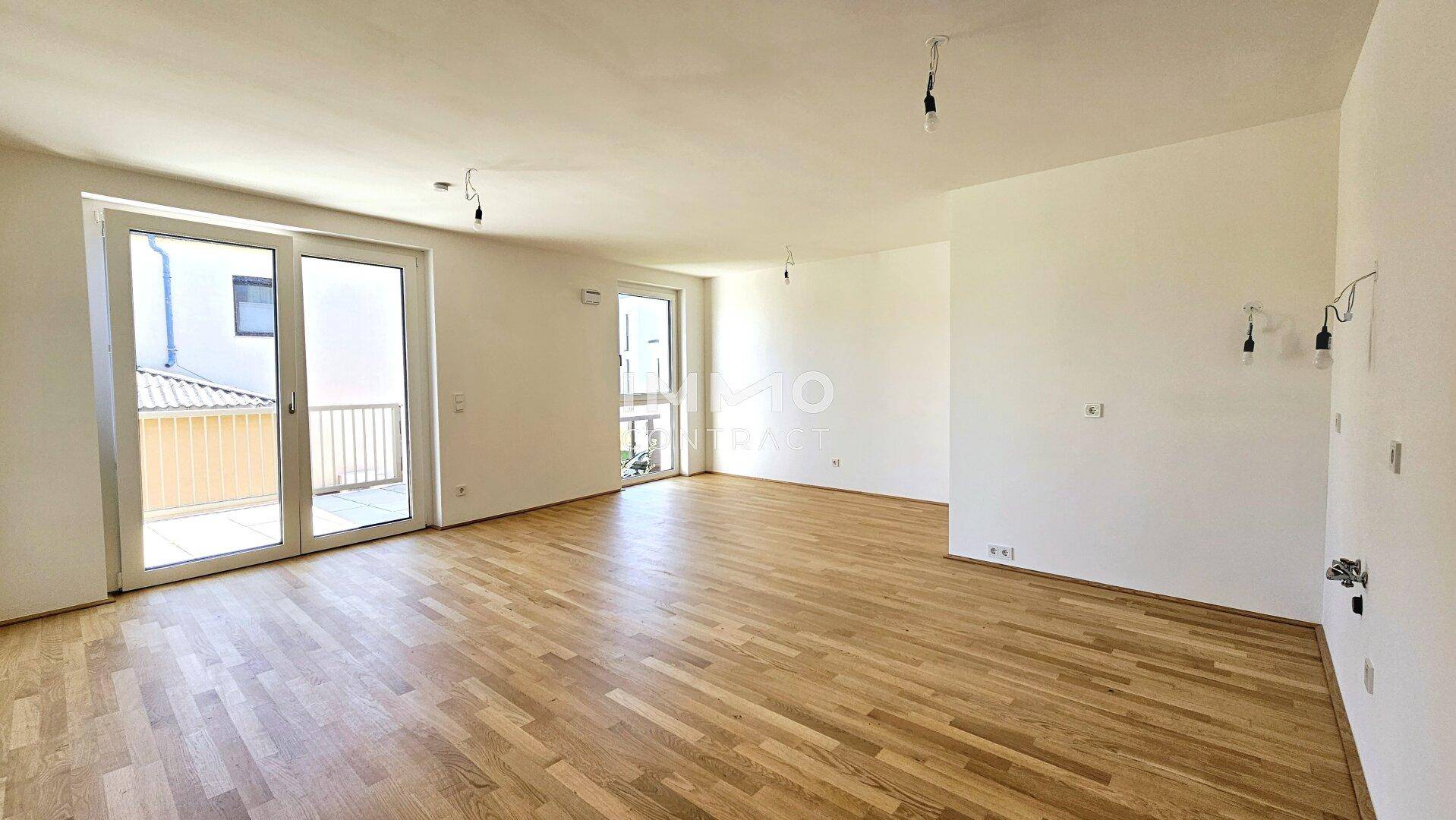 Geräumiges, unmöbliertes Wohnzimmer mit hellem Parkettboden und mehreren Fenstertüren.