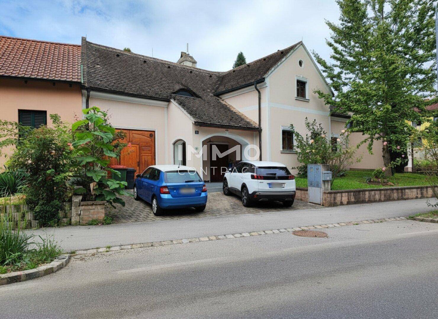 Ansicht des Hauses von der Straße mit Einfahrt und geparkten Autos, umgeben von Grünflächen.