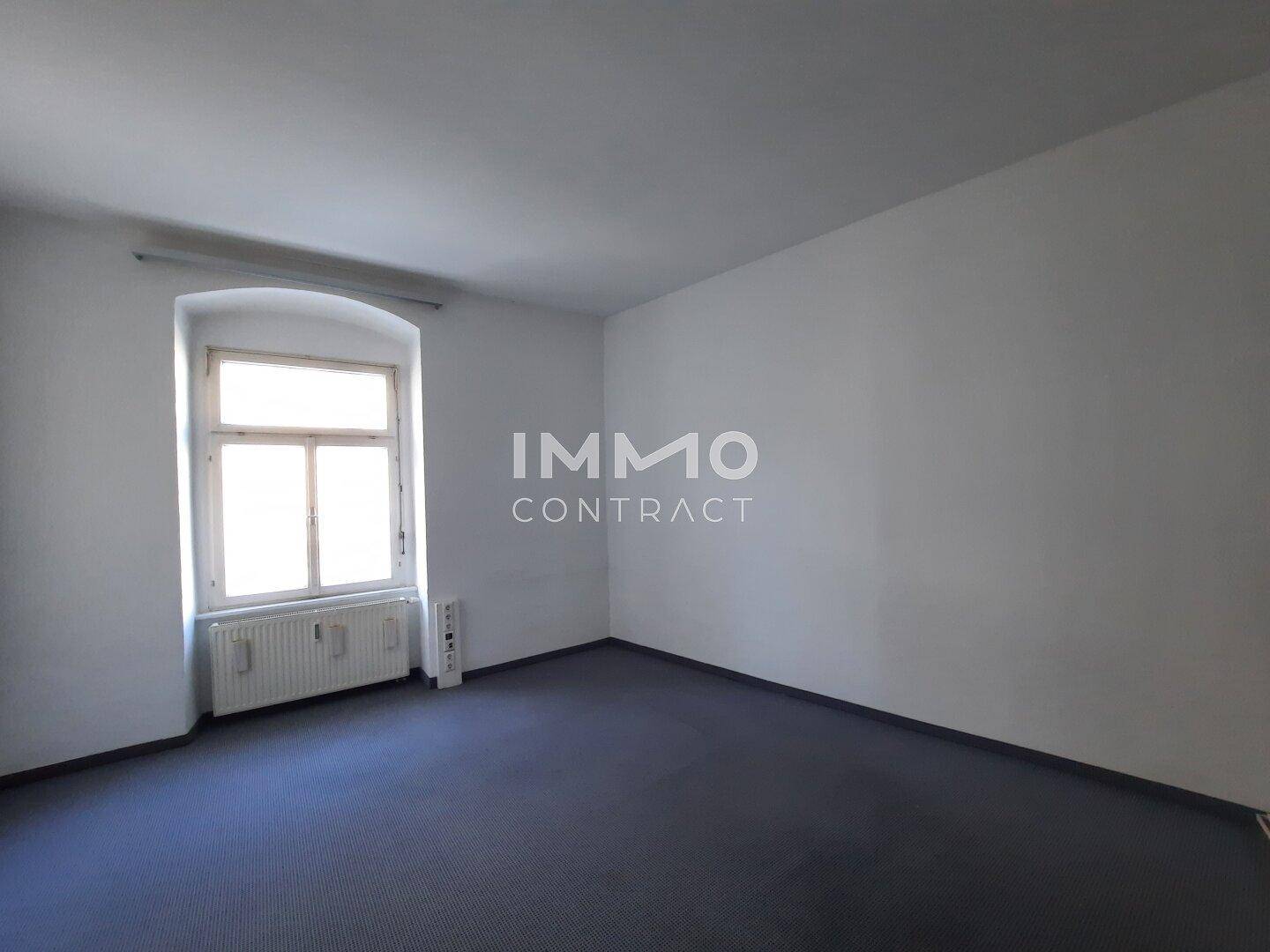 Leerer Raum mit blauem Teppichboden, weißen Wänden und einem Fenster, das viel Tageslicht hereinlässt.
