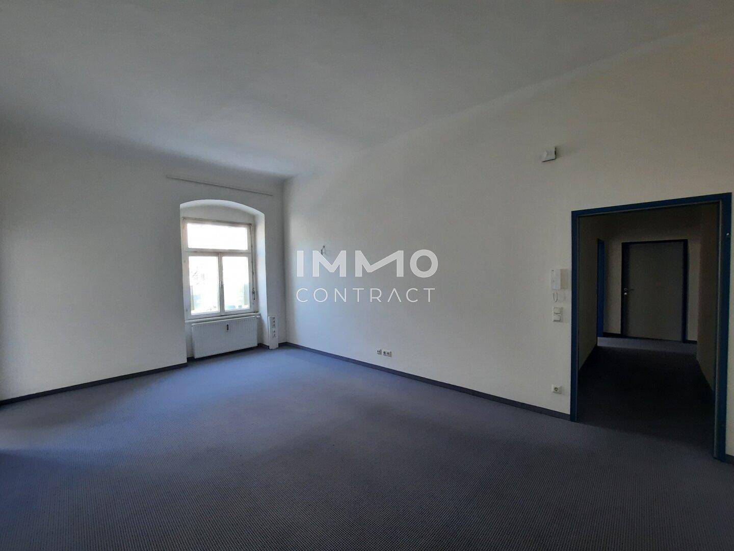 Geräumiger, leerer Raum mit blauem Teppichboden, weißen Wänden und einem Fenster, das Tageslicht hereinlässt.