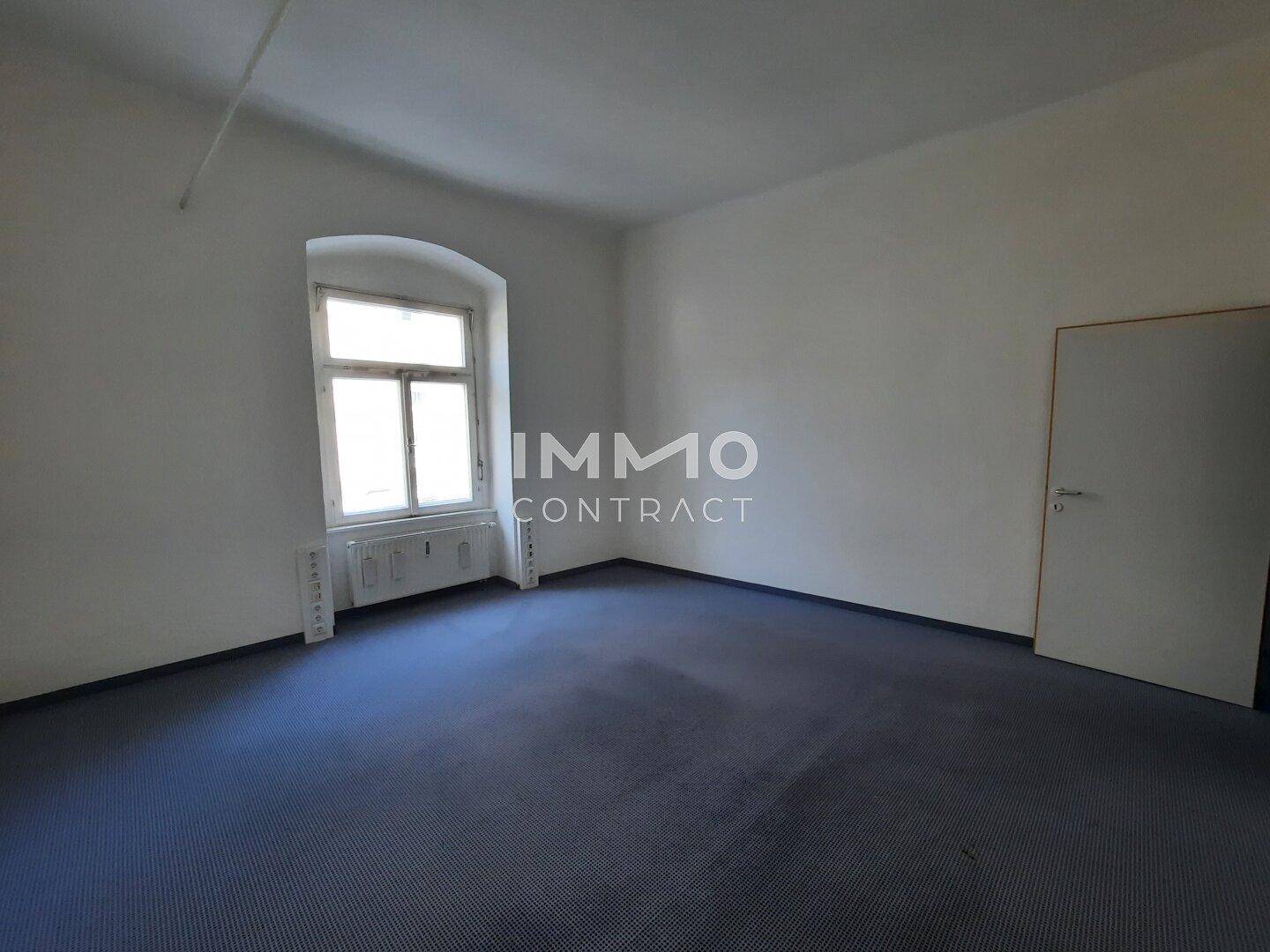 Heller Raum mit blauem Teppichboden, weißen Wänden und einem Fenster mit Heizkörper darunter.