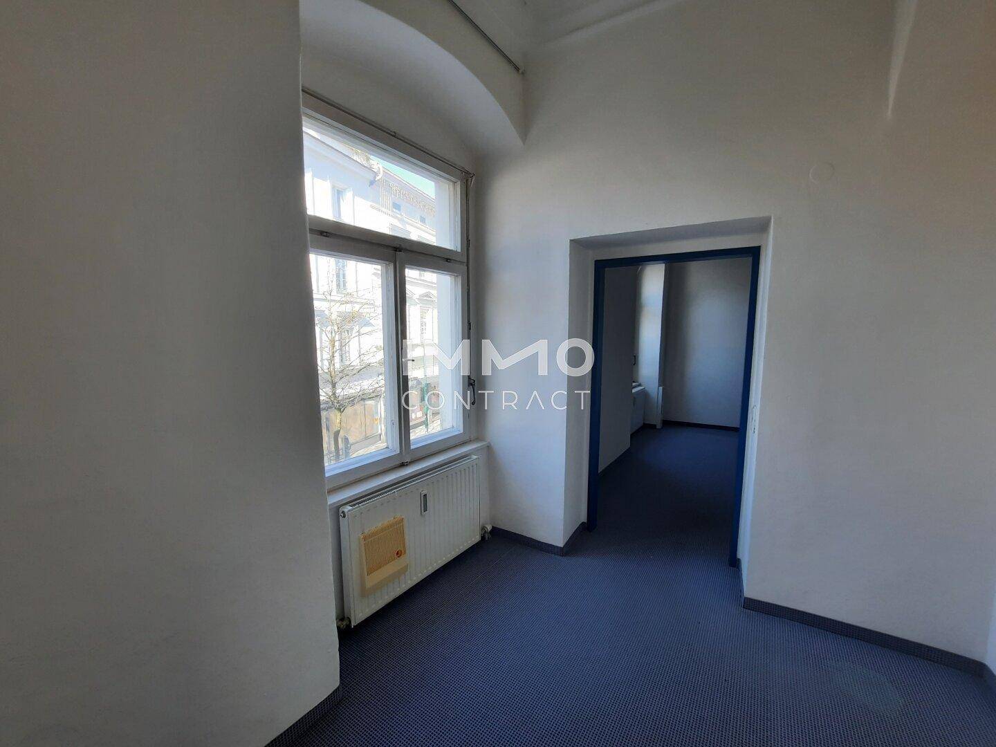 Heller Flur mit großem Fenster, blauem Teppichboden und Blick auf die umliegende Straße.
