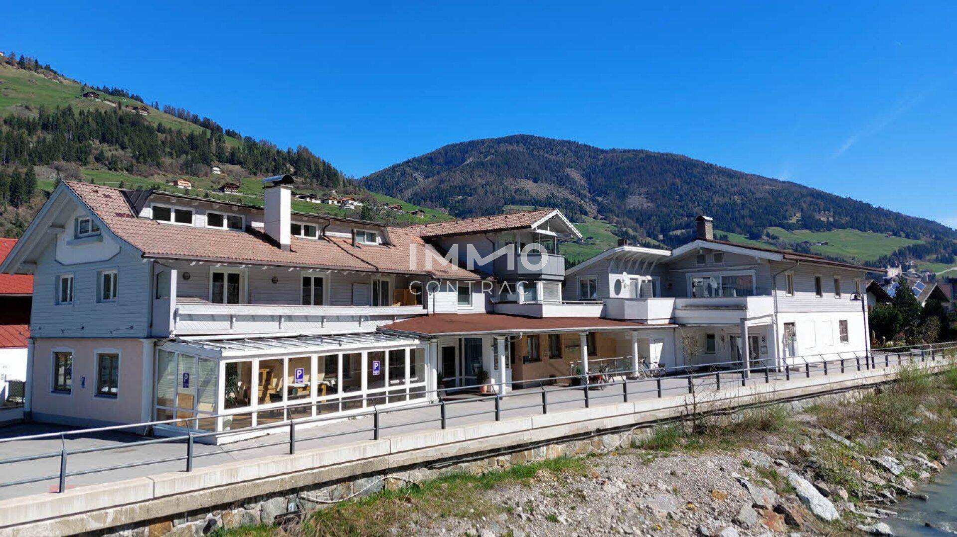 Panoramablick auf den Gebäudekomplex mit angrenzendem Fluss und grünen Berghängen unter blauem Himmel.
