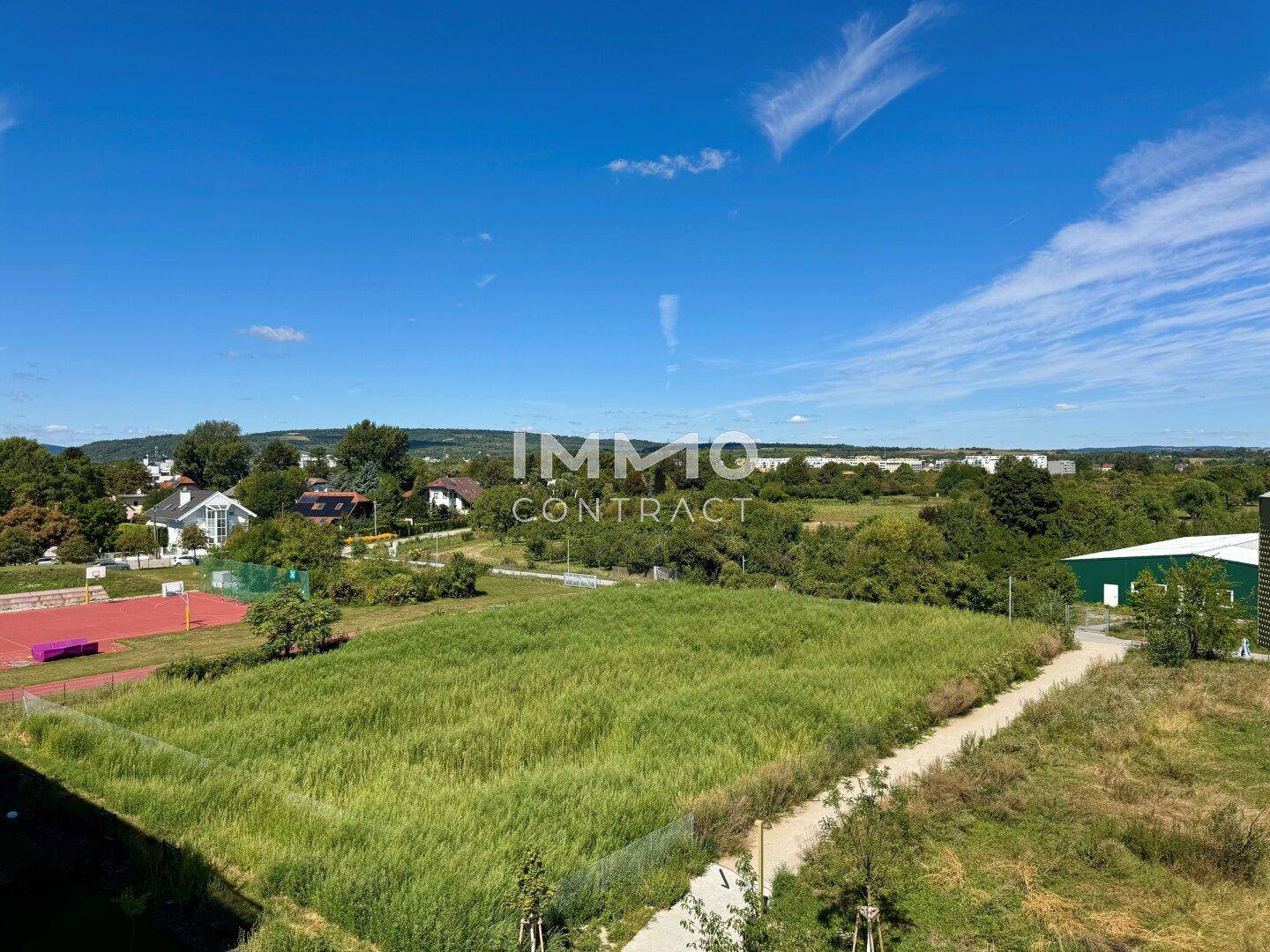 Panoramablick über weite Wiesen und Wohngebiete mit Sportplatz in der Ferne.