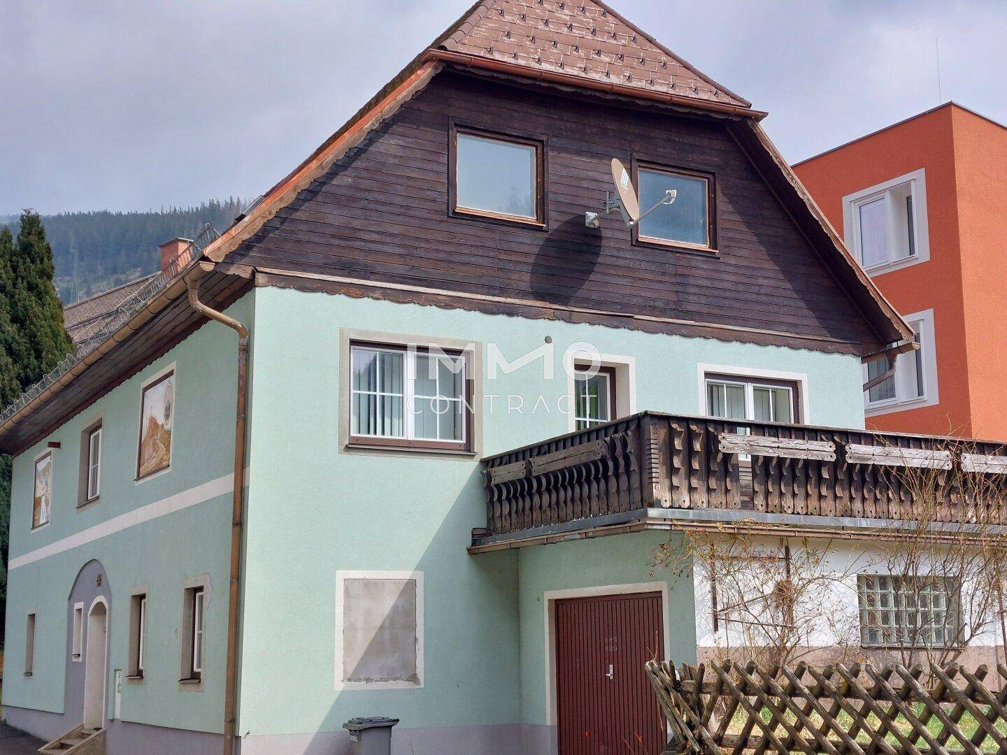 Seitenansicht des Wohnhauses mit traditionellem Holzbalkon und Garage, umgeben von Natur.