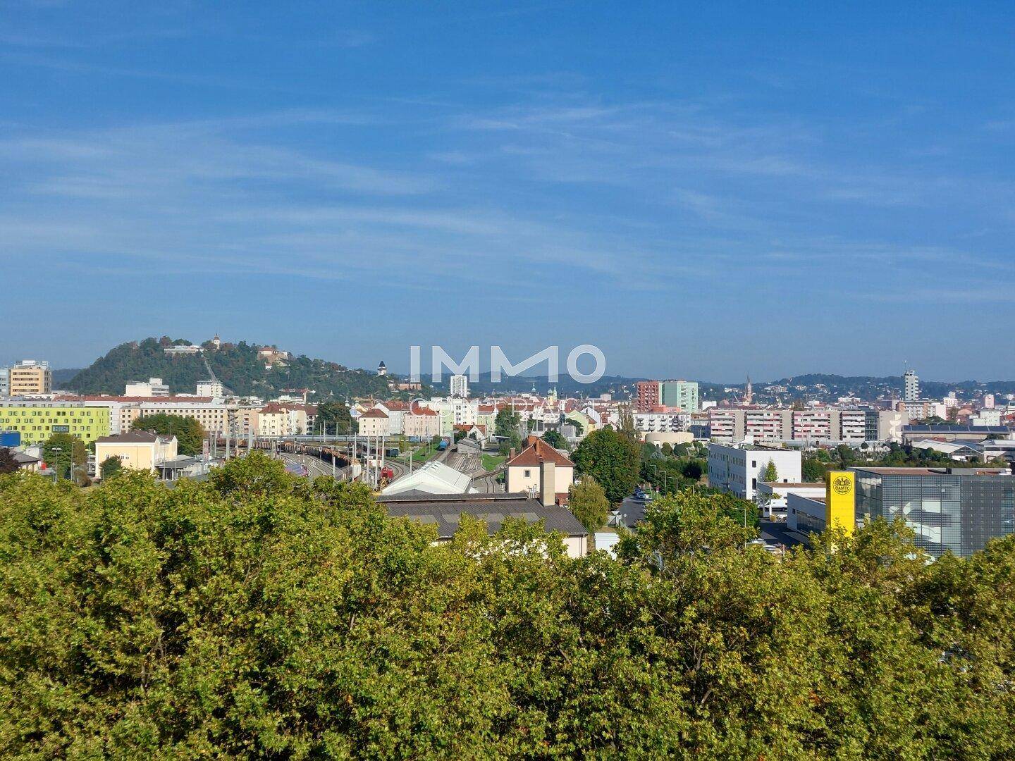 Panoramablick über eine Stadt mit grünen Baumwipfeln im Vordergrund und Hügeln im Hintergrund.