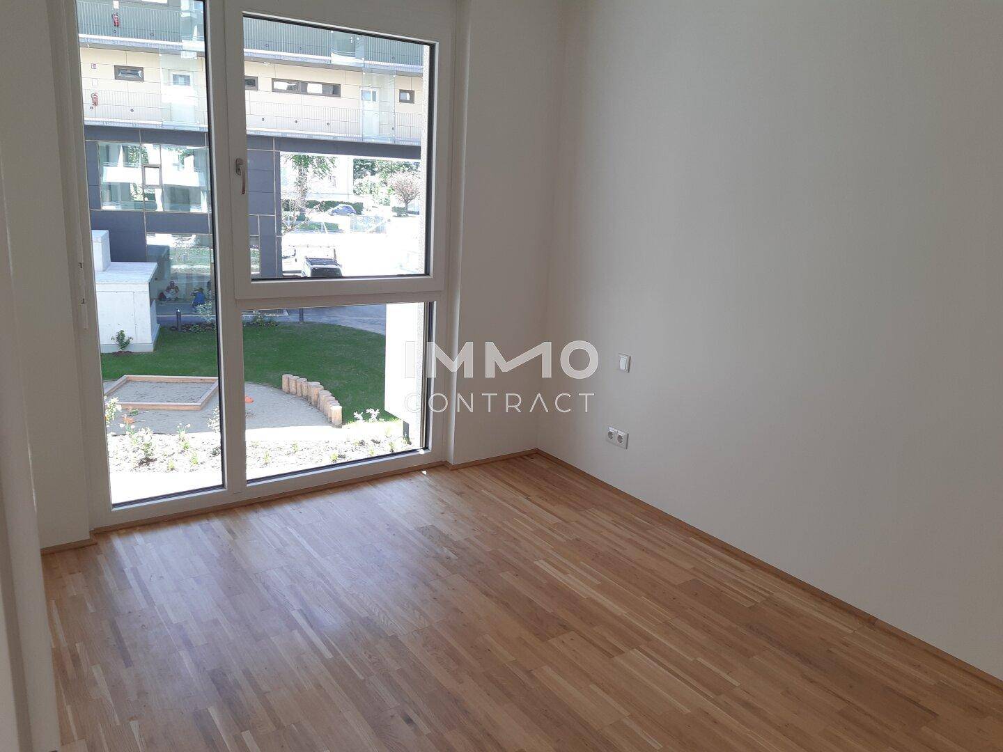 Helles Zimmer mit Parkettboden und großem Fenster, das einen Blick auf einen Sandkasten bietet.