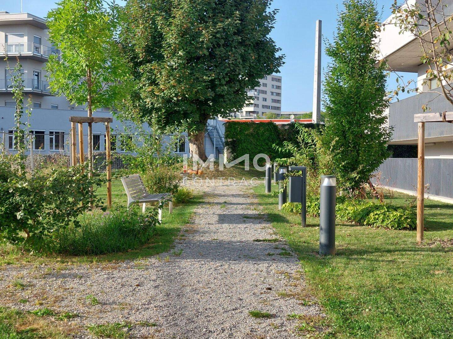 Kiesweg führt durch eine grüne Anlage mit Bäumen und einer Sitzbank im Freien.