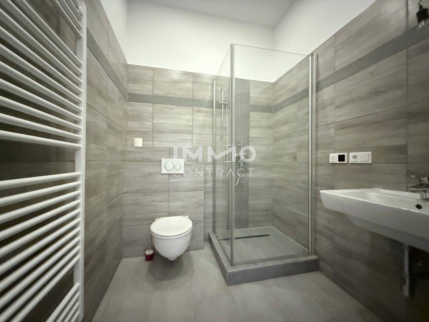 Modernes Badezimmer mit grauen Fliesen, ebenerdiger Dusche, WC und beheiztem Handtuchhalter.