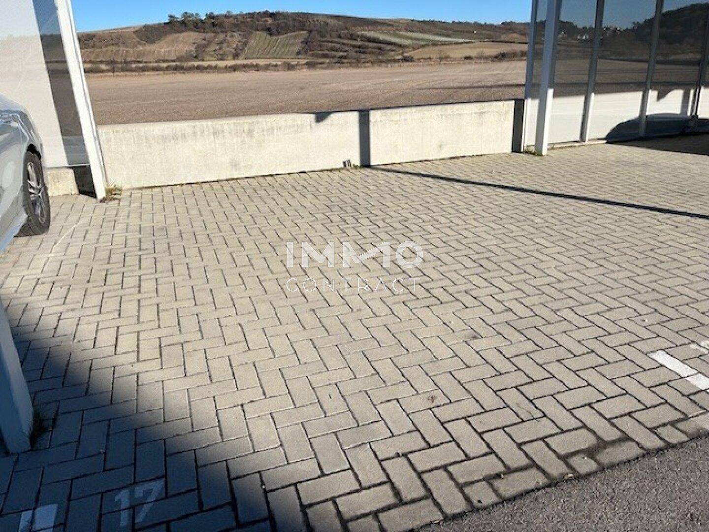 Überdachter Parkplatz mit Pflastersteinen und einer angrenzenden offenen Landschaft im Hintergrund.