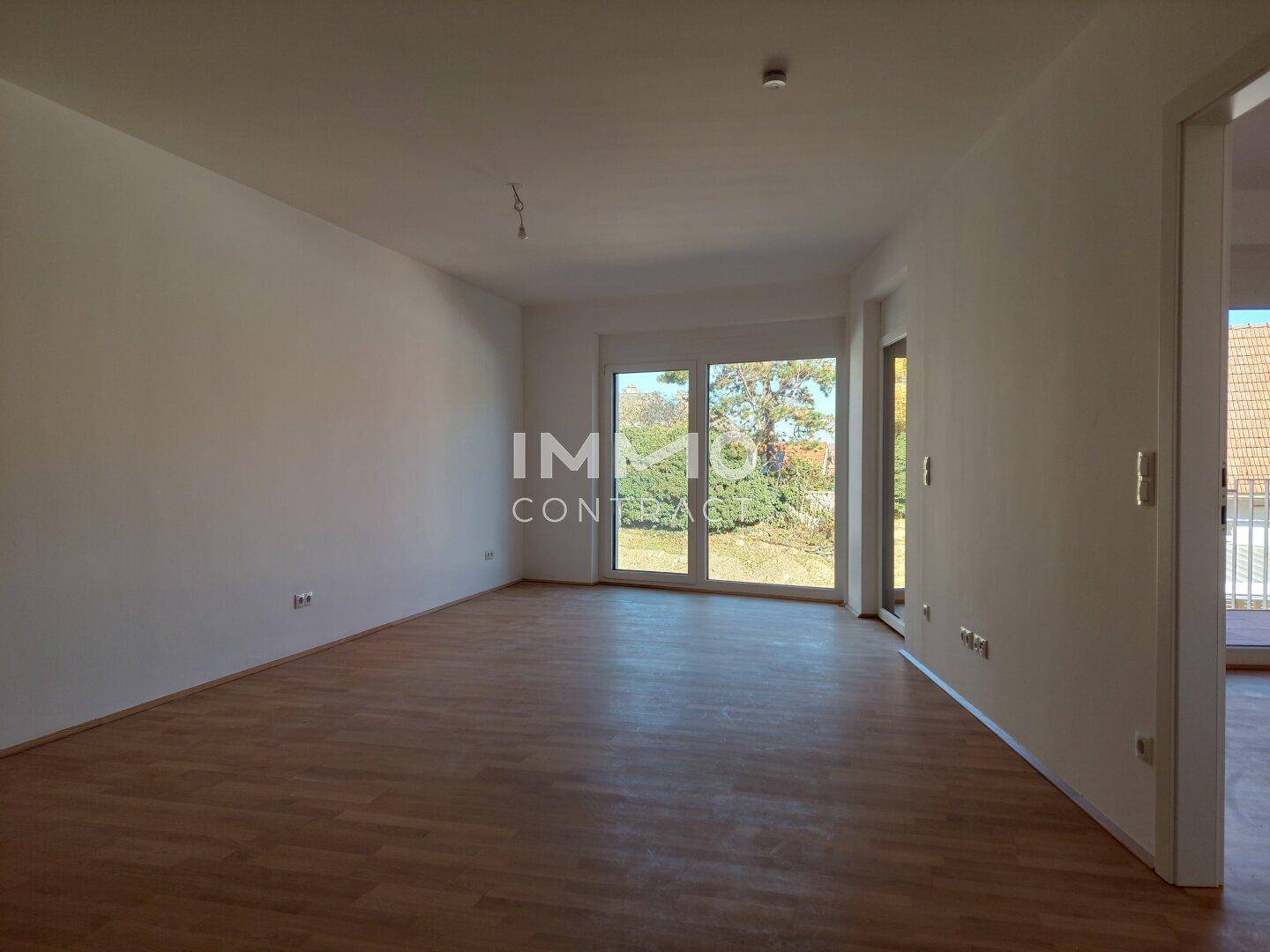 Geräumiges, leeres Zimmer mit Parkettboden und großem Fenster mit Blick ins Grüne.