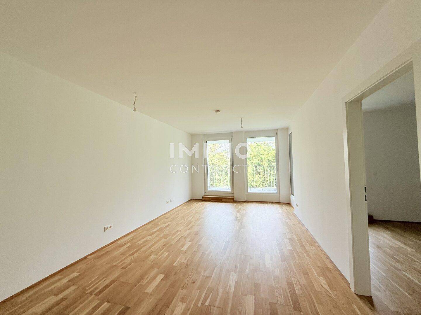 Helles, leeres Wohnzimmer mit Parkettboden und großen Fenstern zum Balkon.