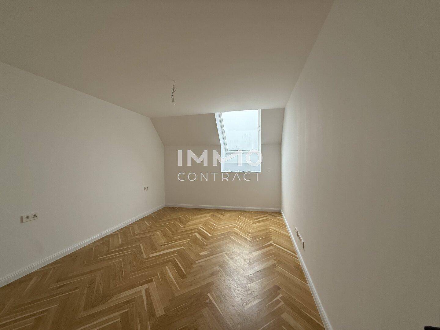 Helles Zimmer mit Fischgrätparkett, schräger Decke und einem Dachfenster für natürliches Licht.