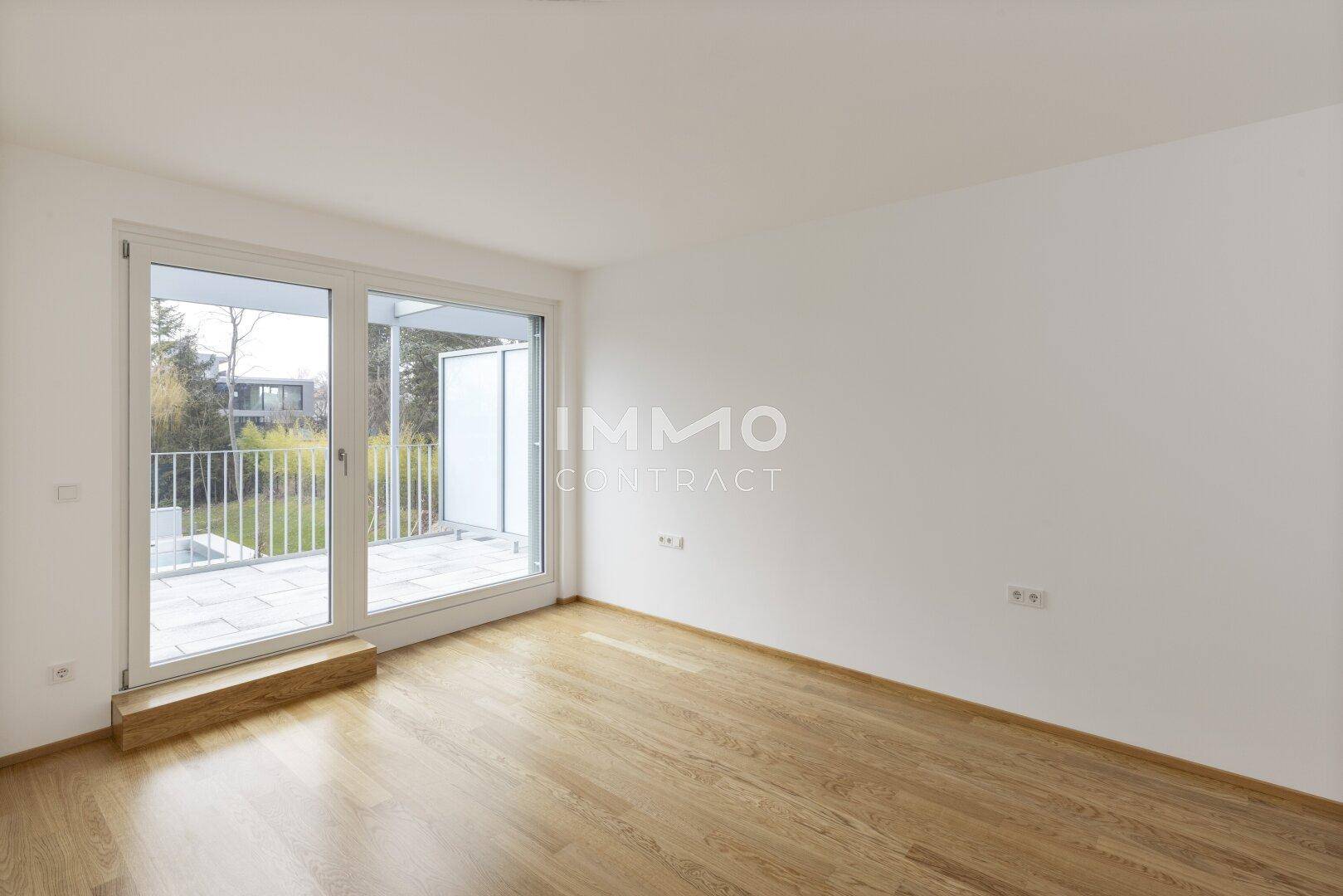 Heller Raum mit Holzboden und großen Fenstern, die Zugang zu einem Außenbereich bieten.
