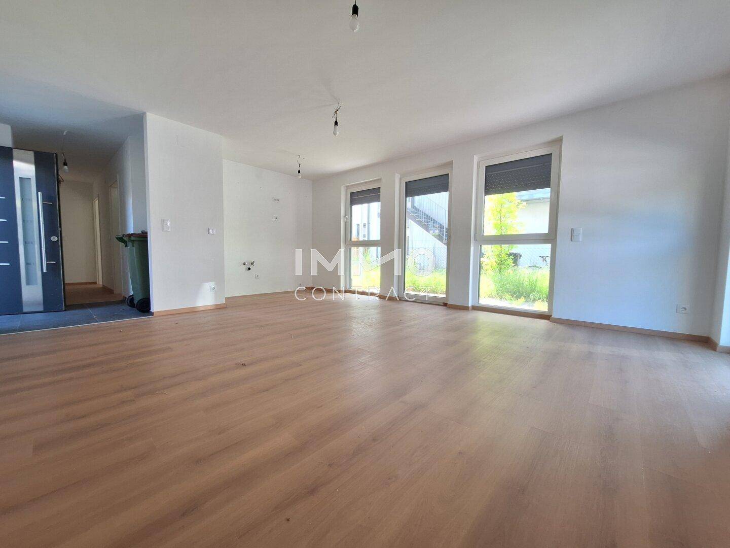 Geräumiges, leeres Wohnzimmer mit hellem Holzboden und großen Fenstern, die viel Tageslicht hereinlassen.