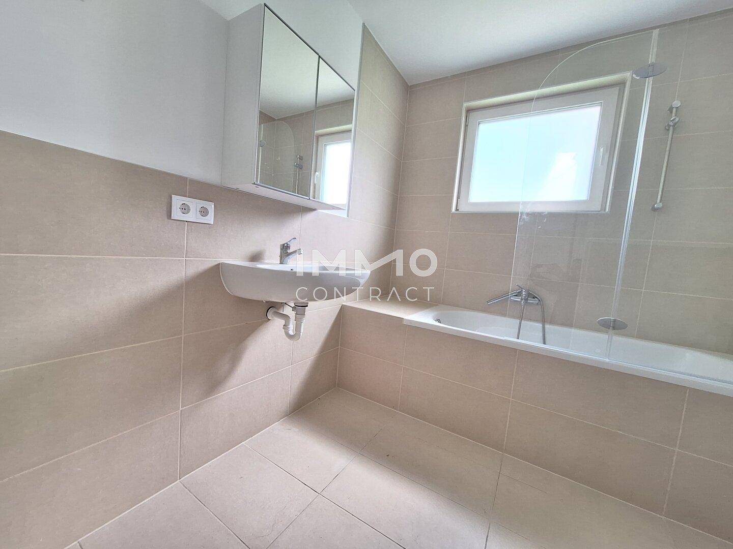 Modernes Badezimmer mit hellen Fliesen, einer Badewanne mit Duschfunktion und einem Fenster.