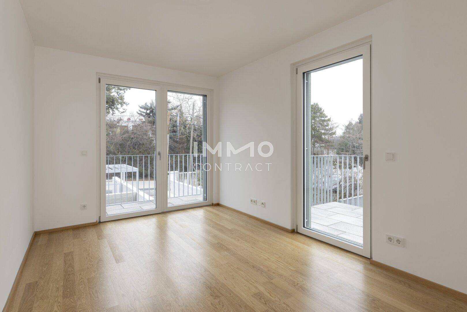 Helles Schlafzimmer mit Holzboden und zwei großen Fenstertüren, die auf einen Balkon führen.