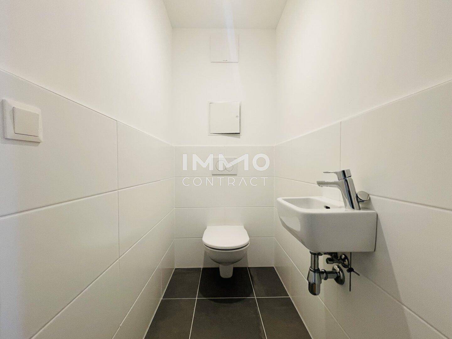 Gäste-WC mit weißen Wandfliesen, dunklem Boden, wandhängender Toilette und kleinem Waschbecken.