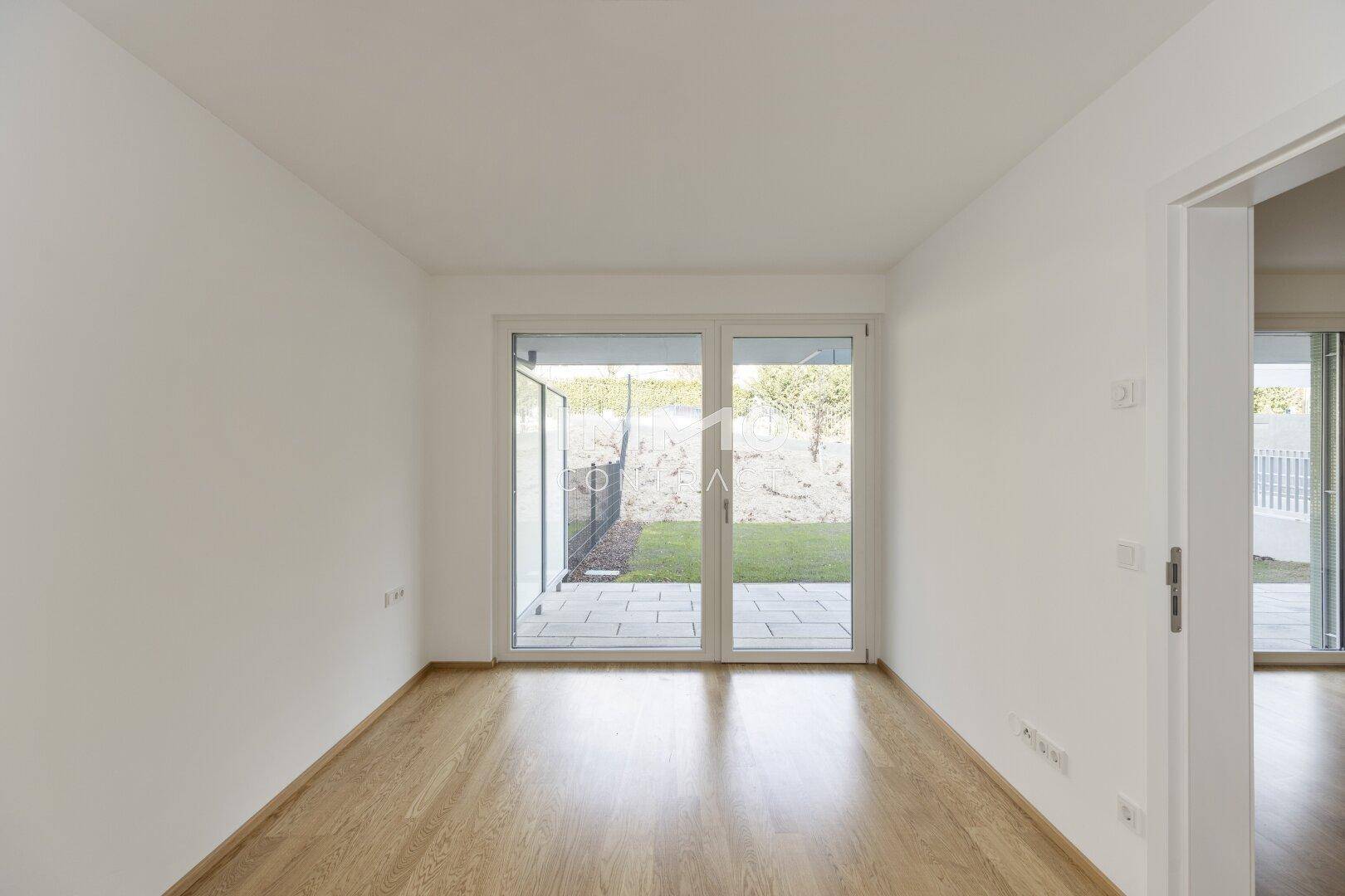 Helles Zimmer mit Holzboden und großen Fenstern, die Zugang zum Garten bieten.