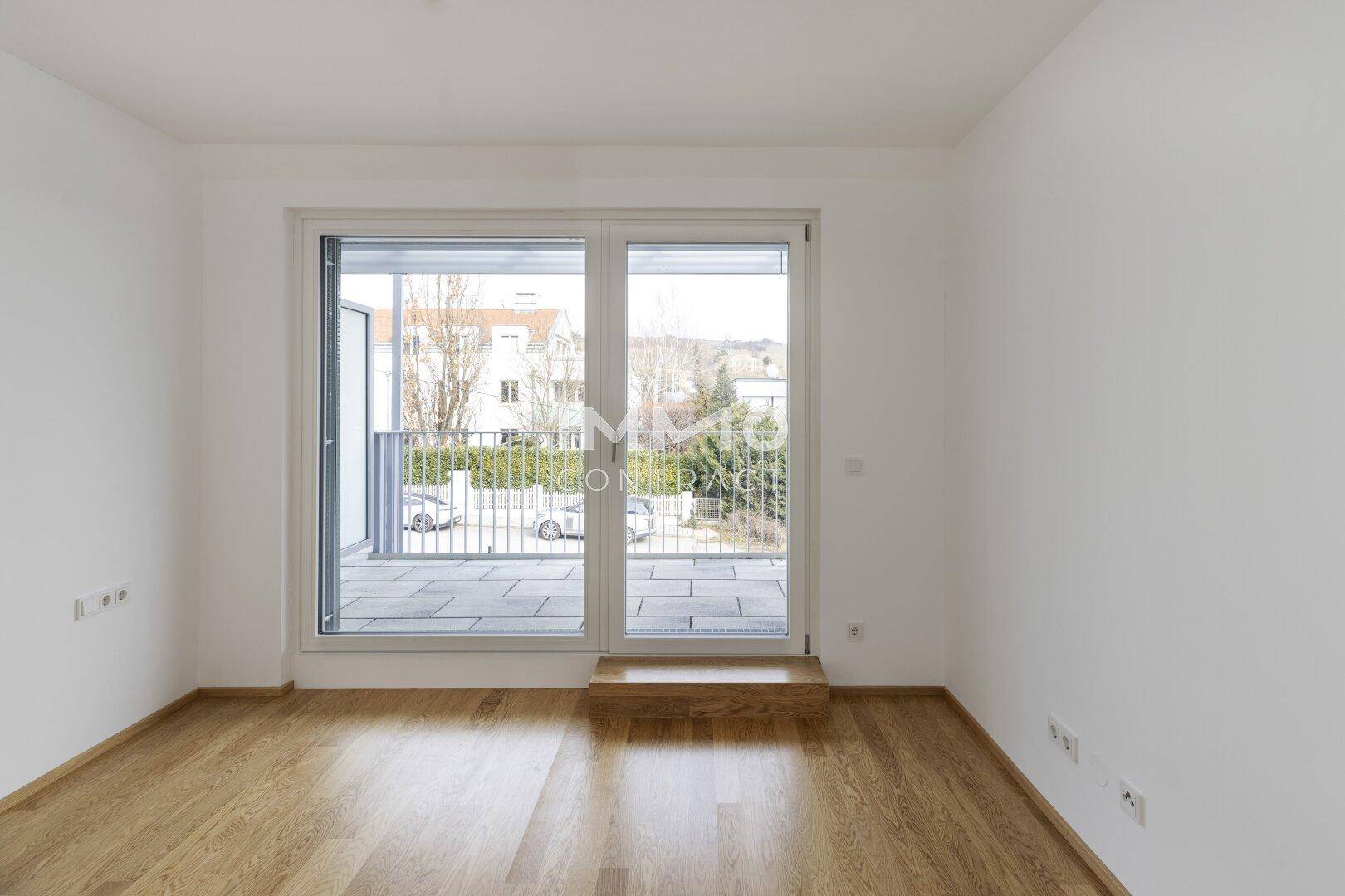 Helles Zimmer mit Holzboden und großem Fenster, das den Blick auf die Straße freigibt.