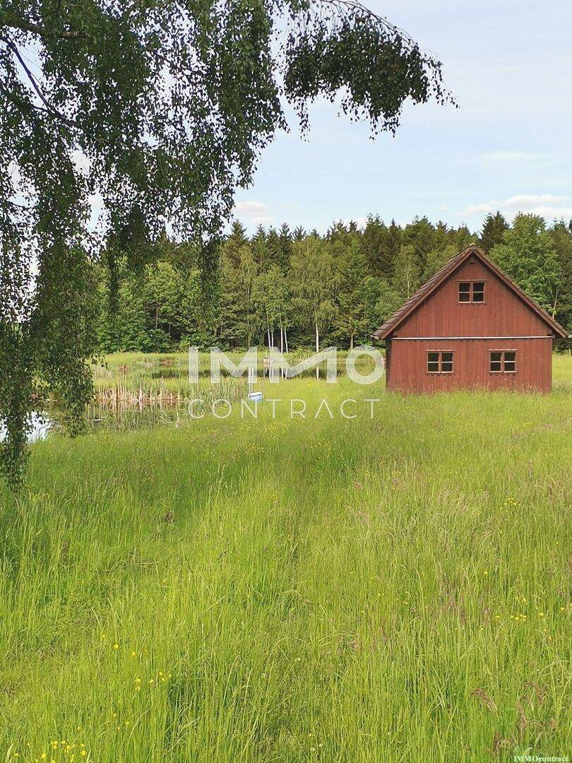 Rustikales rotes Holzhaus in einer weiten grünen Wiese, umgeben von Bäumen und Natur.