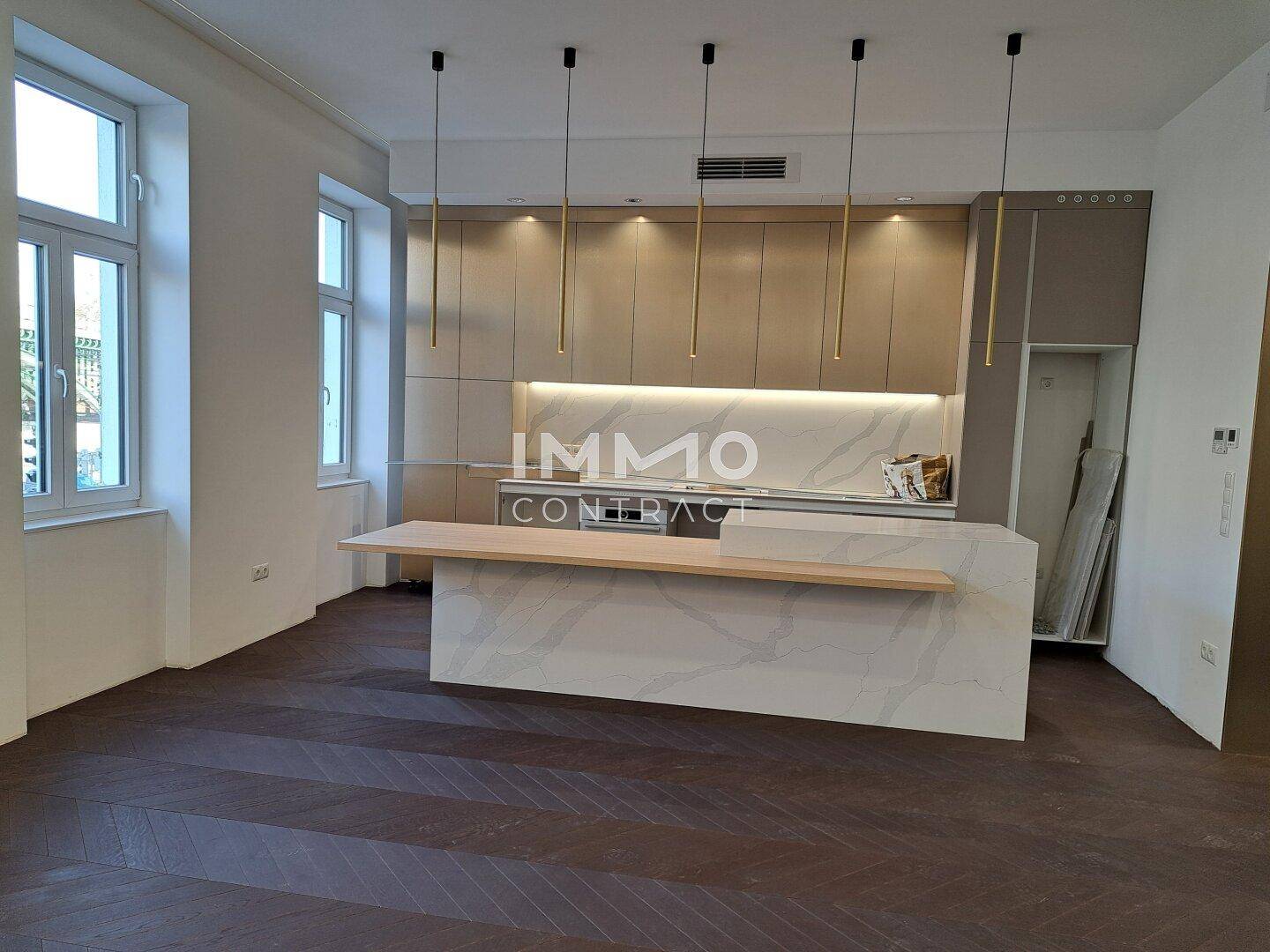Offener Wohn-Essbereich mit moderner Einbauküche, dunklem Fischgrätparkett und eleganten Hängelampen.