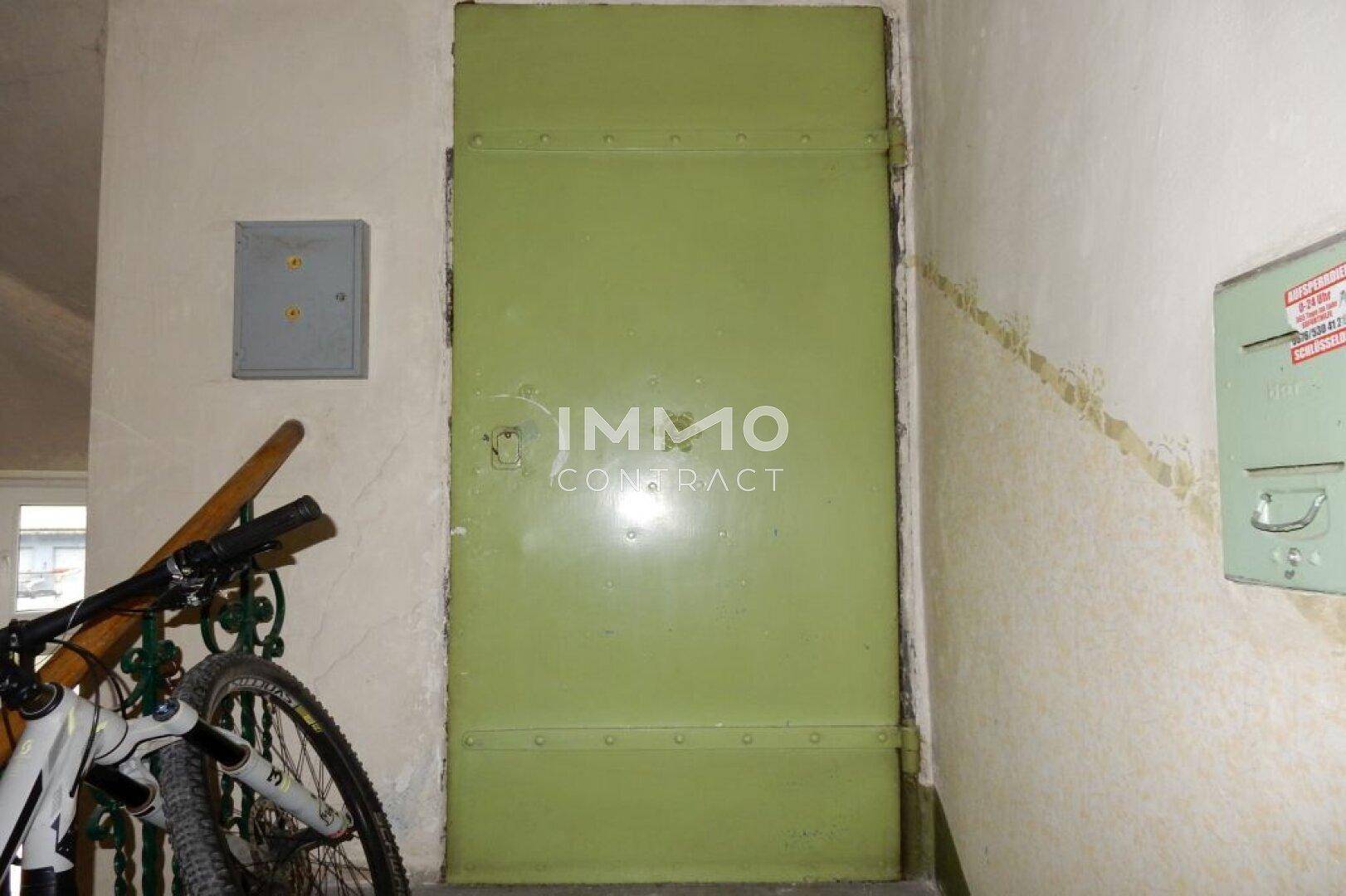 Grüne Eingangstür in einem Flur mit Fahrrad und sichtbaren Gebrauchsspuren an den Wänden.
