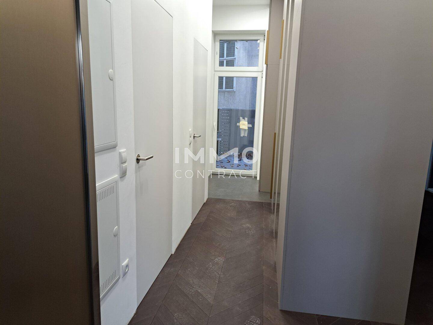 Langer Flur mit dunklem Fischgrätparkett und weißen Wänden, der zu einem Fenster am Ende führt.