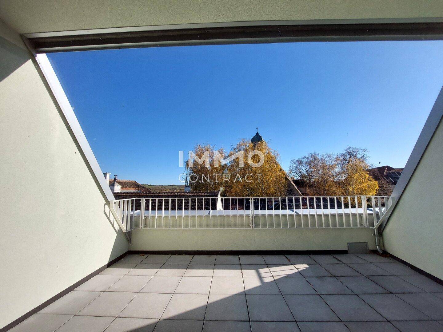 Geräumige geflieste Terrasse mit weißem Geländer und Blick auf einen Kirchturm und herbstliche Bäume.