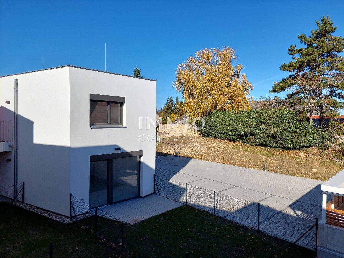 Modernes Gebäude mit kleiner Terrasse und eingezäuntem Gartenbereich, umgeben von Grün.